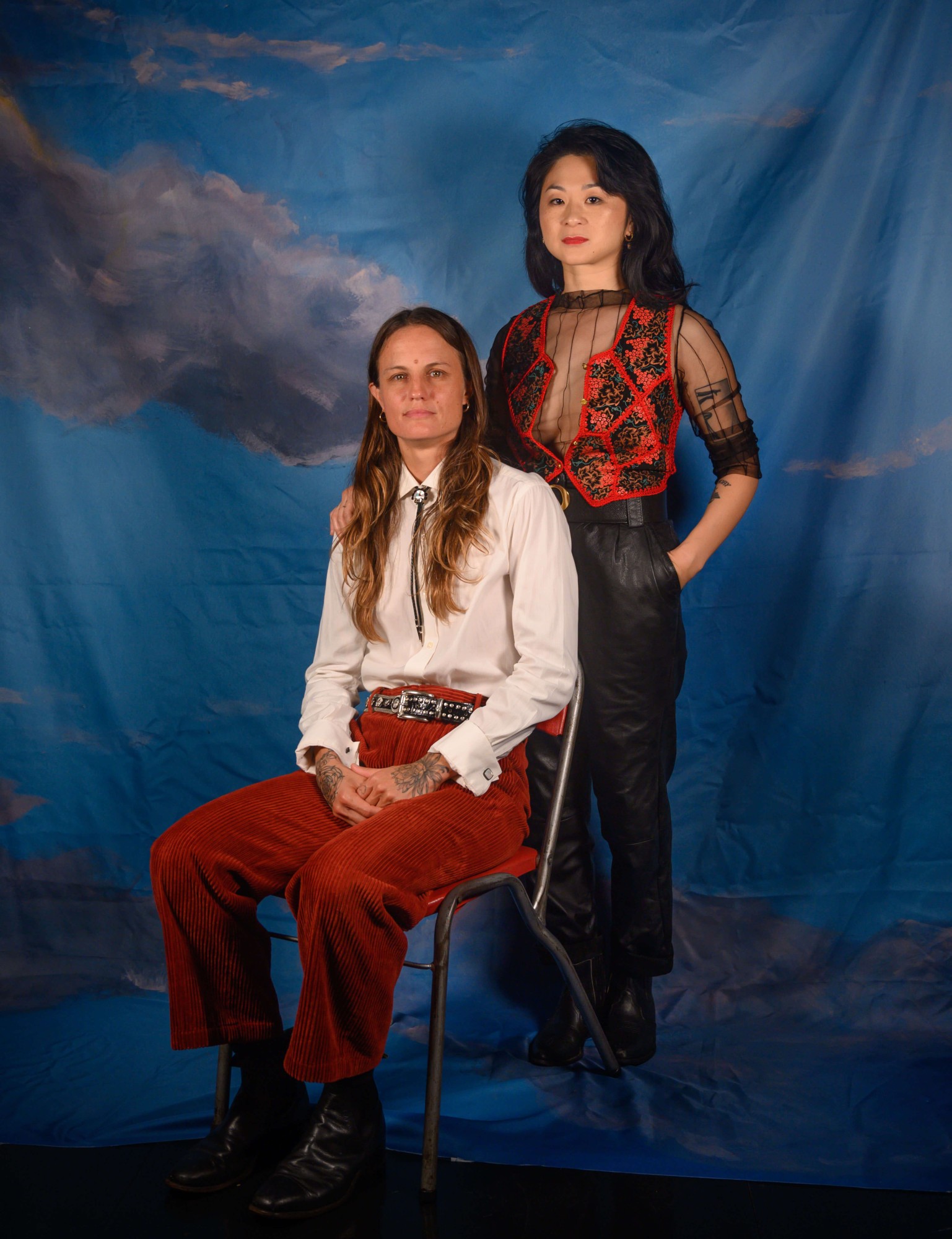 Joy, an Asian person dressed in a embroidered red vest, places her hand on her girlfriend Lyndsay’s shoulder.