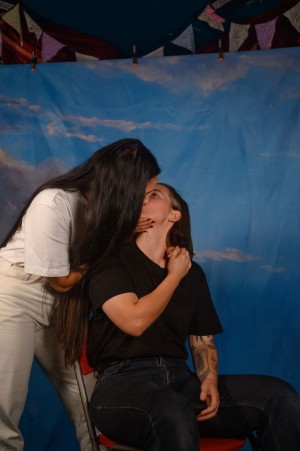 Andy, obscured by her waist length hair, leans down to kiss Kels, who is seated.
