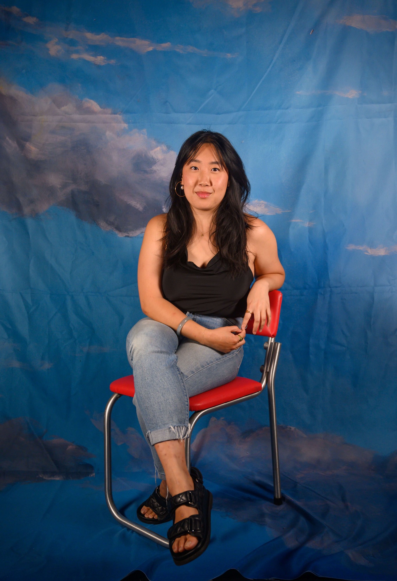Jess, an Asian person, sits relaxed against a red chair.