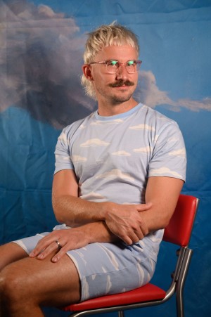 Trent smiles at the camera. He wears a blue co-ord outfit with a white cloud pattern.